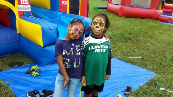 Kids at a party with their faces painted, standing in front of moon bounces.