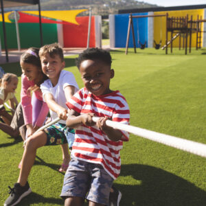 Front view of multi ethnic group of happy school kids playing tug of war in playground