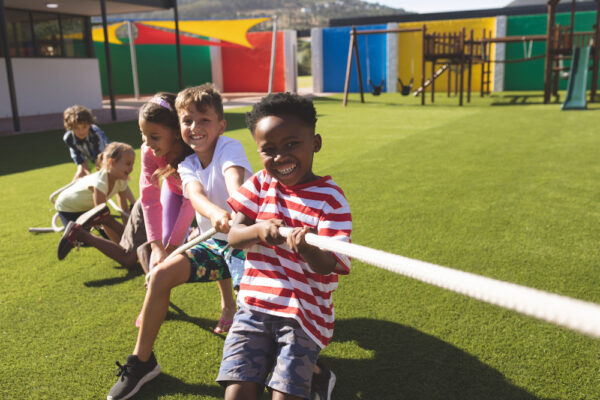 Front view of multi ethnic group of happy school kids playing tug of war in playground